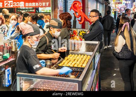 Due cuochi che cucinano takoyaki e polpi al banco da asporto del Museo Dotonbori Konamon, mentre i clienti fanno la fila per i posti all'interno. Notte. Foto Stock