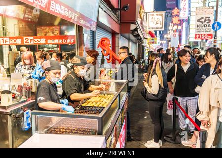 Due cuochi che cucinano takoyaki e polpi al banco da asporto del Museo Dotonbori Konamon, mentre i clienti fanno la fila per i posti all'interno. Notte. Foto Stock