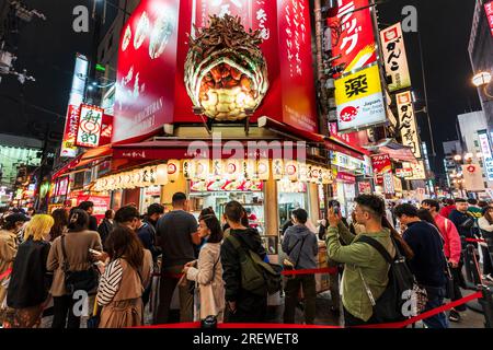 Scena movimentata con persone in coda e a piedi presso il popolare edificio del ristorante Takoyaji Juhachiban in serata a Dotonbori, Osaka. Foto Stock