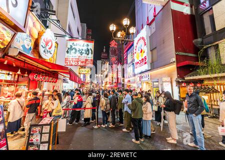 Vista lungo una trafficata e vivace via Dotonbori notturna, Osaka, con la gente in coda al banco take away del ristorante Takoyaki Doraku Wanaka Foto Stock
