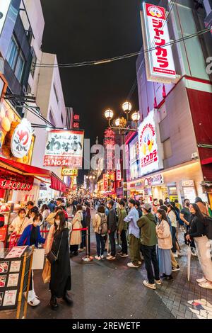 Vista lungo una trafficata e vivace via Dotonbori notturna, Osaka, con la gente in coda al banco take away del ristorante Takoyaki Doraku Wanaka Foto Stock