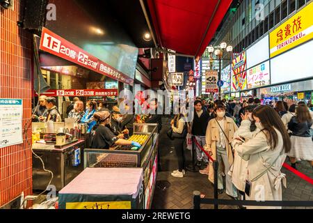 Persone che aspettano fuori dal ristorante del Museo Konamon per i tavoli all'interno la sera a Dotonbori, Osaka. Banco da asporto di fronte all'ingresso. Foto Stock