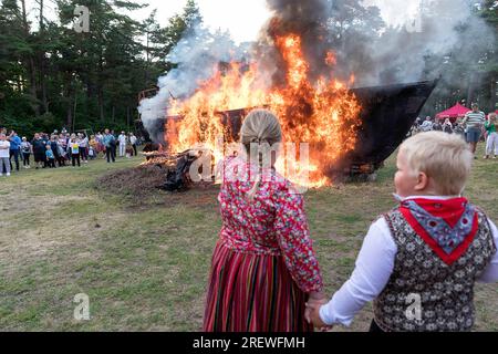 Ragazzo e ragazza locali vestiti con abiti folcloristici estoni che celebrano la jaanipaeva o la giornata di mezza estate con falò (barca a vela in fiamme) sul retro, estonia Foto Stock