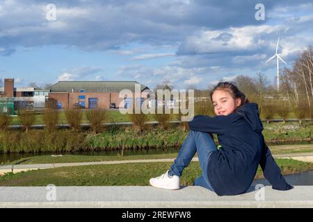 Paesaggio ecologico. Ritratto di una ragazza adolescente sullo sfondo di un cielo tempestoso di primavera e di un mulino a vento elettrico. Foto Stock