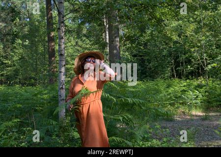 Donna di mezza età in un parco verde ombreggiato. Vestito arancione, cappello di paglia, occhiali da sole. Posiziona per il testo Foto Stock