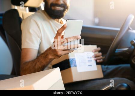 Foto ritagliata del corriere utilizzando il telefono che tiene il pacco in auto Foto Stock