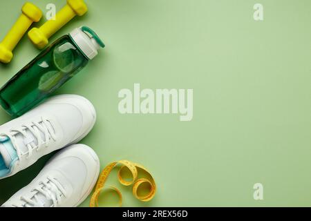 Set creativo di attrezzature sportive e per il fitness. Sneakers bianche da donna, bottiglia per acqua, metro a nastro e manubri verdi su sfondo verde chiaro Foto Stock