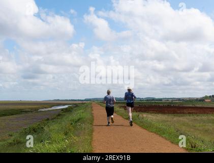 Vista posteriore di due donne che fanno jogging a metà estate lungo un sentiero attraverso le paludi salmastre di Thornham, Norfolk settentrionale, Inghilterra, Regno Unito Foto Stock