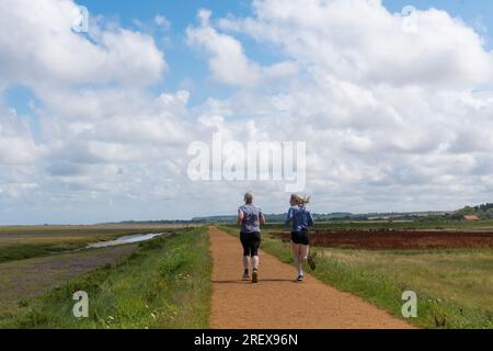 Vista posteriore di due donne che fanno jogging a metà estate lungo un sentiero attraverso le paludi salmastre di Thornham, Norfolk settentrionale, Inghilterra, Regno Unito Foto Stock