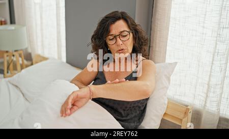 Donna ispanica di mezza età che gratta il braccio per prurito in camera da letto Foto Stock