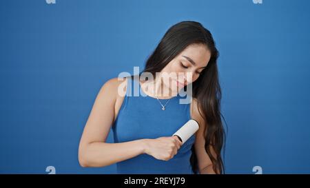 Giovane bella donna ispanica che pulisce la t-shirt con rullo di peli su sfondo blu isolato Foto Stock