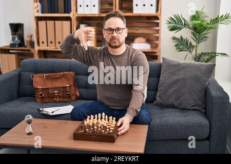 Uomo caucasico di mezza età che gioca a scacchi seduto sul divano con la faccia arrabbiata, segno negativo che mostra antipatia con il pollice abbassato, concetto di rifiuto Foto Stock