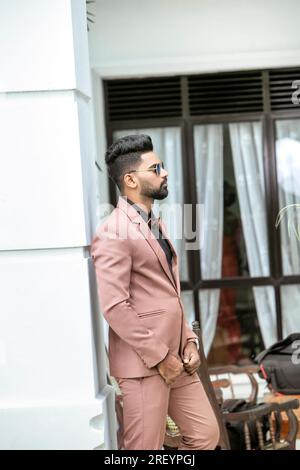 Giovane e affascinante uomo d'affari indiano in costume e occhiali da sole in piedi all'aperto e guardando lontano Foto Stock