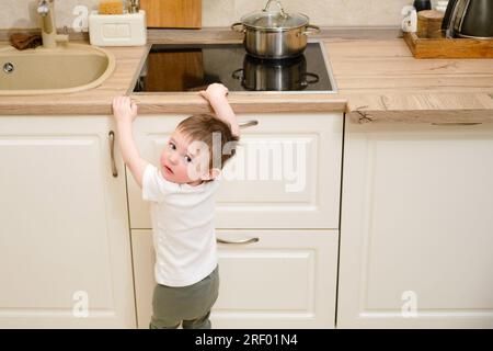 Il bambino salite su una stufa elettrica calda nella cucina domestica. Un bambino piccolo tocca la superficie della stufa con la mano al rischio di ottenere b Foto Stock