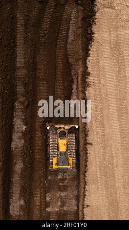 Vista aerea direttamente sopra un bulldozer o una macchina movimento terra con cingoli che spingono terra e terreno nel settore edile in un cantiere in condizioni di terreno brownfield wi Foto Stock