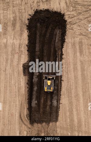 Vista aerea direttamente sopra un bulldozer o una macchina movimento terra con cingoli che spingono terra e terreno nel settore edile in un cantiere in condizioni di terreno brownfield wi Foto Stock