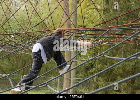 Ragazza adolescente che si arrampica sulla ragnatela con espressione nel parco pubblico. Giorno di sole primaverile Foto Stock