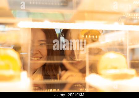 Coppia sorridente asiatica che guarda le torte attraverso il bicchiere nella panetteria e nella caffetteria. Uomo e donna indicano e ordinano dessert mentre si frequentano Foto Stock