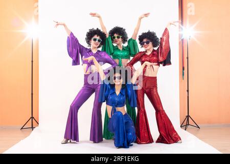 Un gruppo di ragazze con abiti svasati colorati e parrucche afro posa in uno studio fotografico. Stile discoteca degli anni '80 o '70. Foto Stock