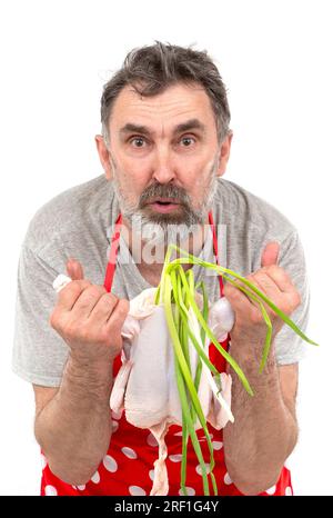 Un anziano cuoco maschio barbuto in grembiule rosso a pois contiene un pollo crudo e un mazzo di cipolle su sfondo bianco. Cuocere con il pollo. Foto Stock