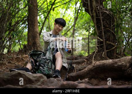 Un escursionista asiatico maschio attivo e felice siede su una roccia, con acqua potabile, mentre riposa durante l'escursione nella foresta. Foto Stock