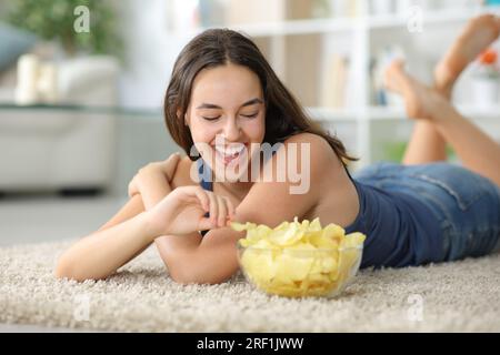 Donna felice che ride e mangia patatine su un tappeto a casa Foto Stock
