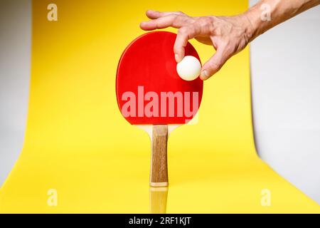 Mano maschile che tiene la palla da ping pong sul lato rosso di una racchetta da ping pong su sfondo giallo. Concetto di gara di ping pong da ping pong. Foto Stock