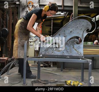 27 luglio 2023, Sassonia, Hohenprießnitz: La fabbro e metal designer Marika Widdermann lavora su una panchina a due posti nella fucina di suo padre Roger Widdermann. Con la loro fucina, il team padre-figlia è una delle poche imprese artigianali che ancora praticano questo antico mestiere in Germania. Oltre ai singoli oggetti utilitaristici e gioielli, producono anche mobili forgiati per i clienti nazionali e stranieri, nonché ciotole e palle da fuoco per il riciclaggio della cera residua, che sono particolarmente richiesti durante i mesi estivi. Foto: Waltraud Grubitzsch/dpa Foto Stock