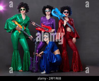 Un gruppo di musica pop canta in un nightclub. Ragazze vestite con costumi retrò degli anni Settanta e divertenti parrucche afro posano insieme. Foto Stock