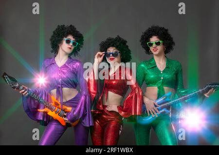 Bellissime ragazze vestite in stile discoteca con parrucche afro posano con le chitarre ai raggi della luce del palcoscenico. Gruppo disco vintage per donne. Foto Stock