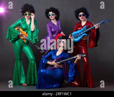 Un gruppo di musica pop canta in un nightclub. Ragazze vestite con costumi retrò degli anni Settanta e divertenti parrucche afro posano insieme. Donne felici che si divertono a h Foto Stock