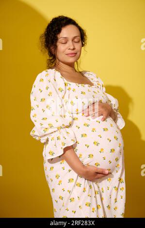 Felice donna incinta, gravida madre in attesa con elegante abito da sole bianco estivo, che tocca la sua grande pancia, posa con gli occhi chiusi, godendo il suo pregno Foto Stock