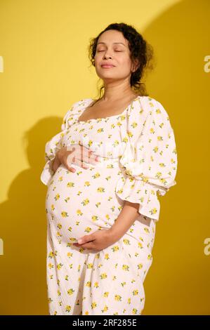 Felice donna incinta, bella madre in attesa che mette le mani sulla pancia, si diverte a calci, isolata su sfondo giallo. Gravidanza e madre Foto Stock