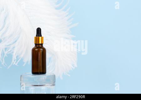 Prodotto cosmetologico per la cura del viso. Siero in flacone di vetro con pipetta su podio di vetro e piuma di struzzo bianco grande su sfondo blu. Essenziale Foto Stock