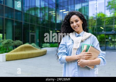 Ritratto di una giovane e bella studentessa ispanica, donna fuori da un campus universitario che sorride e guarda la macchina fotografica, tiene libri di lavoro, libri e libri di testo per studiare. Foto Stock