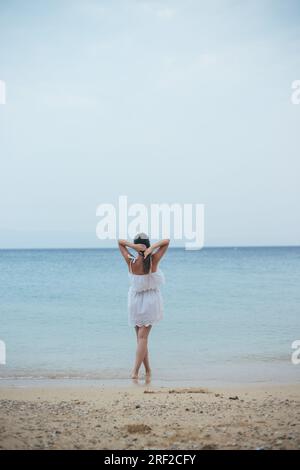 Vista posteriore di una donna in abito bianco estivo in piedi sulla spiaggia sabbiosa Foto Stock