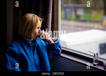 Una donna beve caffè e fuma cannabis. Una donna adulta di più di quaranta anni è seduta vicino alla finestra, accanto ad essa è un dispositivo per fumare marijuana. Foto Stock