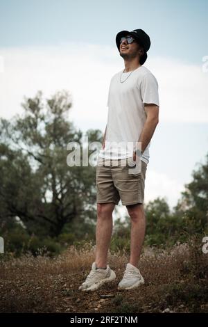 Un uomo di vent'anni, che ride apertamente con una t-shirt bianca, pantaloncini beige, occhiali da sole e un berretto da marinaio nero. È circondato dalla bellezza naturale di Foto Stock