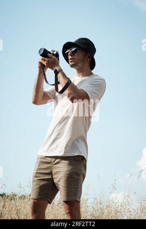 Un ritratto di un uomo di vent'anni, vestito casualmente con una t-shirt bianca, pantaloncini beige, occhiali da sole e un berretto da marinaio nero, con una classica fotocamera slun Foto Stock