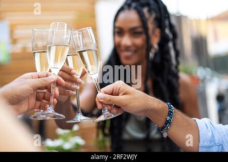 Nel cortile di un ristorante, quattro flauti di champagne si incontrano in un brindisi tra amici. Una donna afro-americana sorridente con i capelli intrecciati è visibile, unendosi Foto Stock