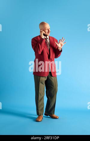 Foto a tutta lunghezza. Uomo emozionale con baffi, vestito in stile vintage in piedi e che parla su un telefono retrò su sfondo blu. Foto Stock