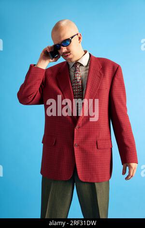 Ritratto di un uomo calvo con baffi, vestito in stile vintage in piedi e che parla su un telefono retrò su sfondo blu. Foto Stock
