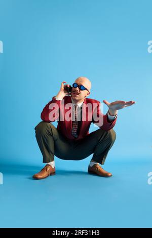 Uomo dalla piega con baffi indossato in stile vintage, accovacciato e parla su un telefono retrò guardando lontano su sfondo blu. Foto Stock