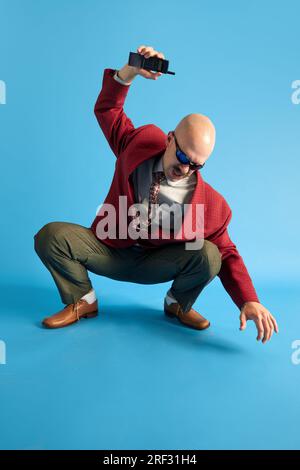 Un uomo arrabbiato con baffi che indossa un'accoccolata in stile vintage per abbattere un telefono retrò su uno sfondo blu da studio. Foto Stock