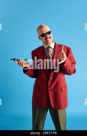 Ritratto di un uomo calvo sorridente con baffi vestito da una giacca retrò rossa con moquette vintage in mano su sfondo blu da studio. Foto Stock
