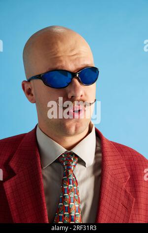Colpo di testa di fischi uomo calvo con baffi vestito da abito retrò business che si nasconde sullo sfondo blu dello studio. Foto Stock