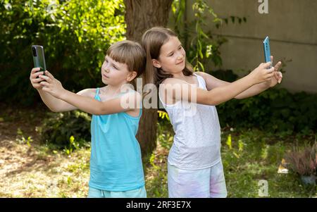 Due bambine che tengono in mano il cellulare del loro smartphone scattano selfie insieme sorridendo, ritratti all'aperto, all'esterno. Bambini e social media t Foto Stock