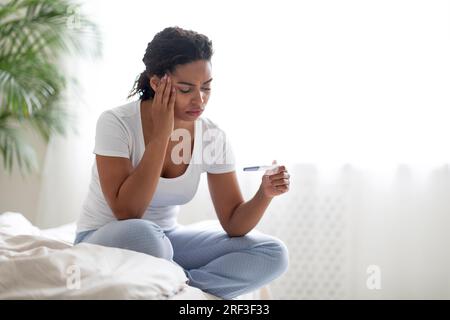 Infelice triste giovane donna afroamericana con test di gravidanza seduta sul letto Foto Stock