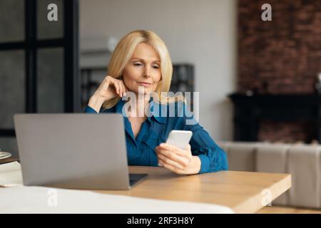 Donna d'affari matura e concentrata che lavora da casa, seduta alla scrivania di fronte a un moderno pc portatile e utilizza il cellulare Foto Stock