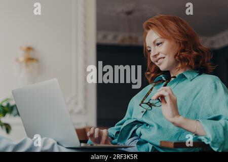 Redhead Woman si è concentrata, felice di lavorare a distanza da un appartamento moderno, preparando una pubblicazione web. Occhiali in mano. Foto Stock